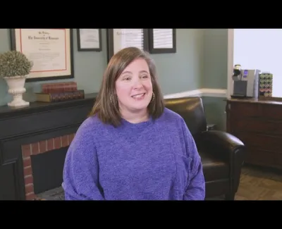 Woman in a purple sweater smiling while seated in a cozy office with framed certificates and a coffee machine.