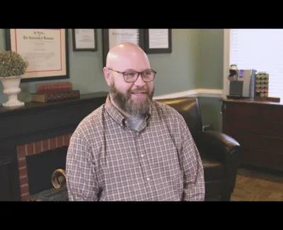 Smiling bearded man wearing glasses and plaid shirt sitting in cozy office with framed certificates and furniture.
