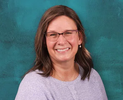 Smiling woman with glasses wearing a light purple sweater against a teal textured background.