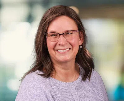 Smiling woman with glasses wearing a light purple sweater in a softly blurred indoor setting.