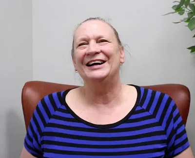 Smiling woman in a blue and black striped shirt sitting in a brown chair next to a green leafy plant indoors.