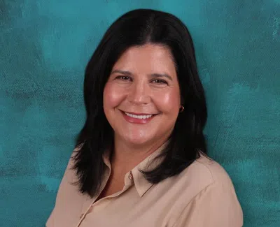 Smiling woman with dark hair wearing beige blouse against a textured teal background