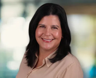 Smiling woman with dark hair wearing a beige blouse in a softly blurred indoor setting.
