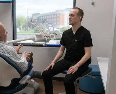 Dentist in black scrubs consulting an elderly male patient seated in a dental chair by a window.