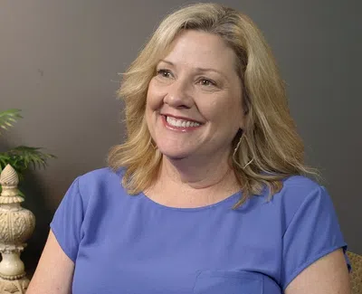 Smiling blonde woman in blue shirt sitting indoors with green plant and decorative ornament in background
