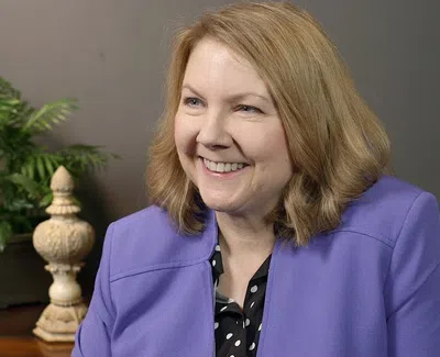 Smiling woman in a purple blazer sitting indoors with plants and decorative items in the background
