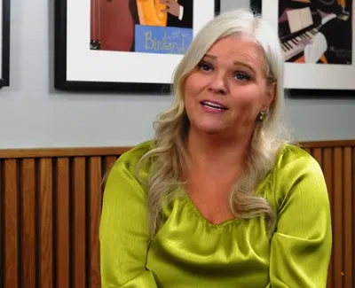 Blonde woman in a green blouse speaking while seated in a room with wooden paneling and framed artwork.