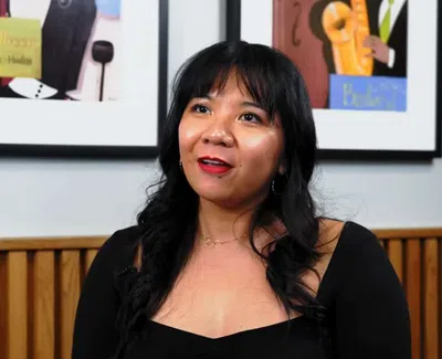Woman with black hair and red lipstick speaking indoors with framed artworks on wall behind her.