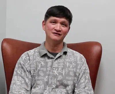 Man with short black hair wearing a patterned gray polo sitting on a brown chair against a plain wall.
