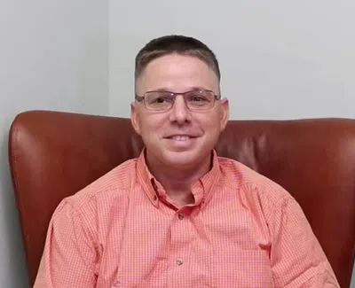 Man wearing glasses and an orange checkered shirt sitting on a brown leather chair against a plain wall.