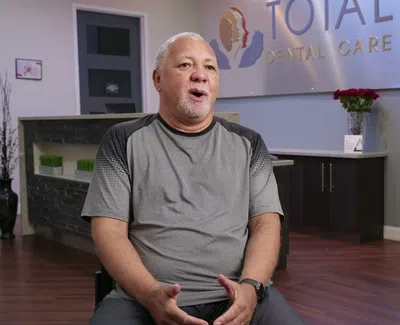 Man sitting and speaking inside a dental clinic with Total Dental Care sign and a smiling woman poster.