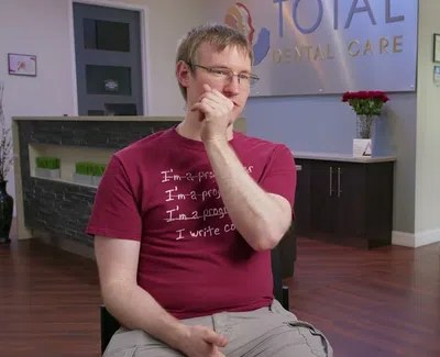 Man in red shirt sitting in a Total Dental Care office with a smiling woman on an Invis banner in the background.