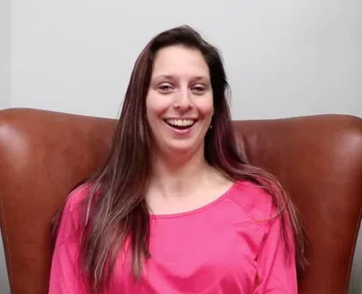Smiling woman with long brown hair wearing a pink shirt sitting on a brown leather chair next to a green leafy plant.