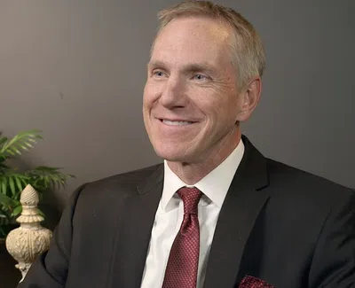 Smiling man in black suit and red tie seated indoors with green plant and dark gray wall background