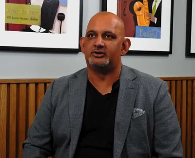 Man in gray blazer and black shirt sitting against wooden paneling and colorful framed artwork background.