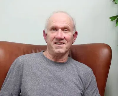 Smiling elderly man in a gray shirt sitting on a brown chair with green leafy plant nearby indoors.