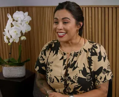 Smiling woman with floral dress sitting indoors near a white orchid plant on a black stand
