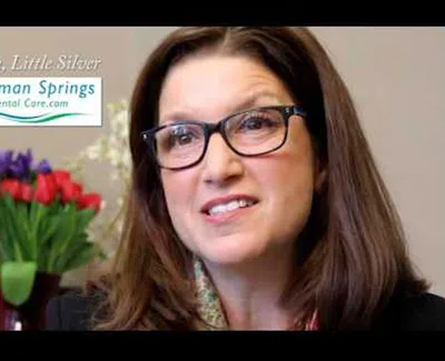 Woman with glasses smiling, Newman Springs Dental Care logo and red and purple flowers in background