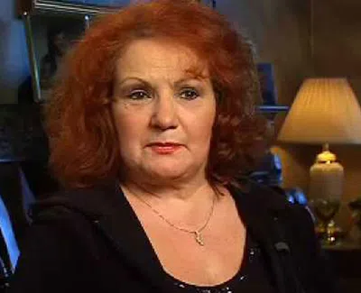 Mature woman with red curly hair wearing a necklace, sitting indoors next to a lit table lamp in a cozy room.