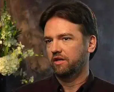 Man with beard speaking in an interview setting with a floral arrangement in the blurred background