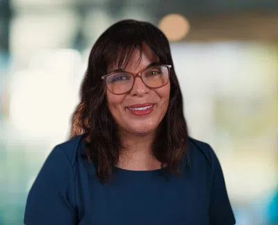 Smiling woman with glasses and dark hair wearing a blue top against a blurred modern office background