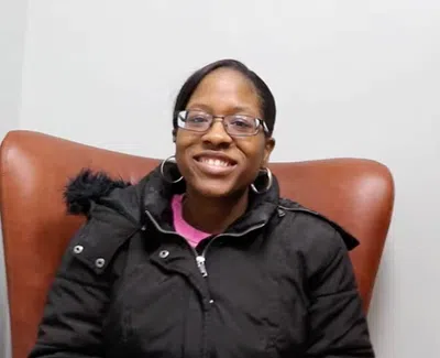 Smiling woman with glasses wearing a black winter coat sitting in a brown chair against a white wall with a green plant.
