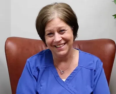 Smiling woman in blue scrubs sitting in brown chair next to a potted green plant against a white wall.