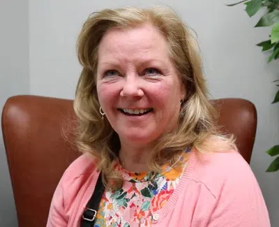 Smiling woman with blonde hair wearing a floral shirt and pink cardigan sitting in a brown chair indoors.
