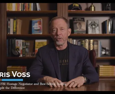 Chris Voss sitting at a desk with bookshelves in the background, identified as former FBI negotiator and author.