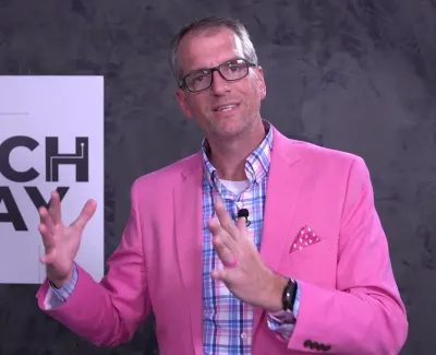 Man in pink blazer speaking in front of Tech Day sign on gray background during presentation