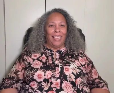 Smiling woman with curly gray hair wearing a floral blouse sitting against a plain wall background