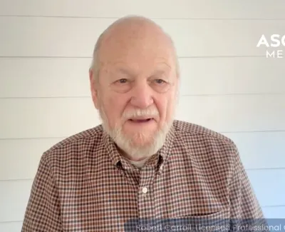 Elderly man in checkered shirt speaking in front of white shiplap wall with Ascend Medical logo