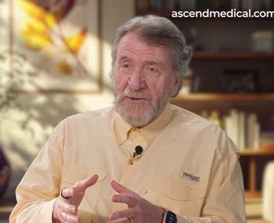 Older man with gray beard speaking in a well-lit room with plants and artwork in the background.