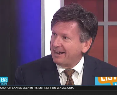 Man in a dark suit and patterned tie speaking on a news talk show with a blue and orange background.