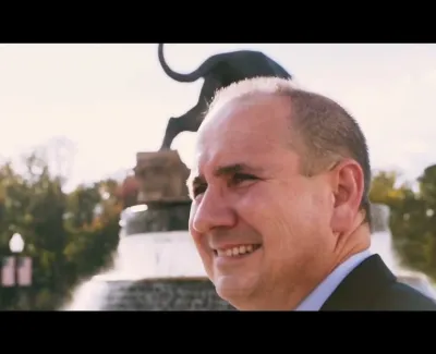 Smiling man in a suit near a water fountain with a black panther statue and trees in the background.