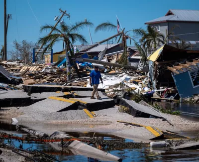 Man walking on broken road amidst hurricane destroyed houses and palm trees under blue sky