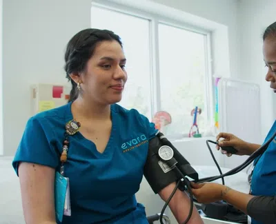 Nurse checking blood pressure of a woman wearing blue scrubs in a bright medical clinic room.
