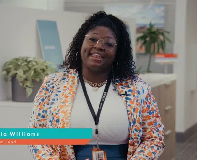 Smiling woman wearing glasses and floral blazer sitting in modern office with badge and lanyard.