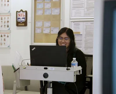 Receptionist smiling while assisting a standing woman at an office desk with laptop and water bottle.