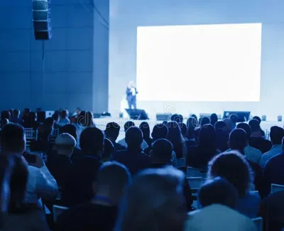 Audience seated in a dimly lit conference room listening to a speaker presenting on a large screen stage.