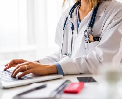 Female doctor in white coat typing on laptop with stethoscope around neck in bright office setting