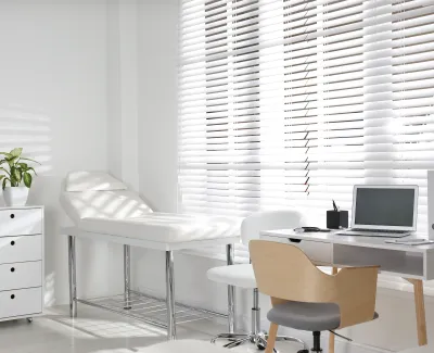 Bright modern medical office with examination table, desk, laptop, microscope, and large window blinds.