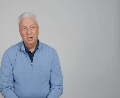 Senior man with white hair wearing a light blue quarter-zip sweater against a plain gray background.