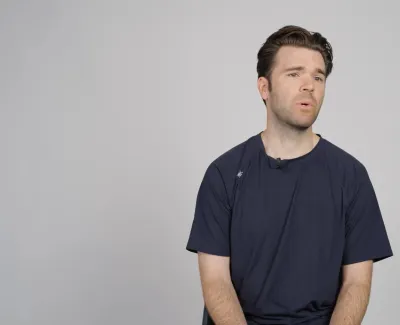Young man in a dark blue t-shirt sitting and speaking against a plain gray background.