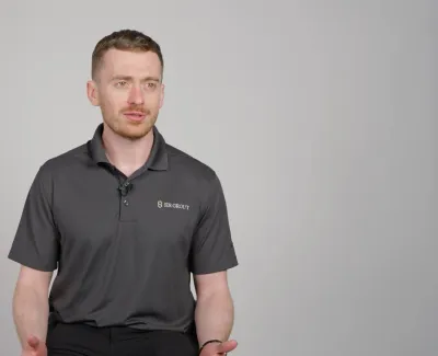 Man wearing a black Sir Grout polo shirt speaking against a plain gray background in a studio.