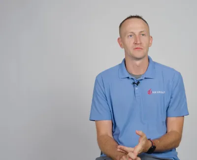 Man in blue company polo speaking against plain gray background with hands clasped and focused expression