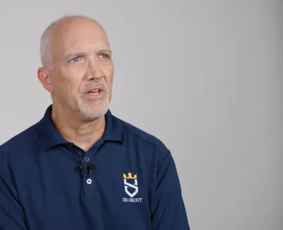 Middle-aged man with gray hair and goatee wearing a navy blue polo shirt with a logo against a plain background