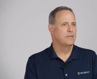 Middle-aged man in navy polo with Sir Grout logo speaking against plain grey backdrop