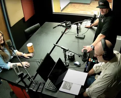 Three people recording a podcast in a soundproof studio with microphones, laptops, and headphones.