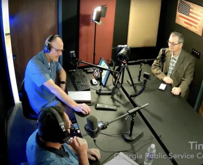 Three men in a podcast studio with microphones, headphones, recording equipment, and an American flag on the wall.